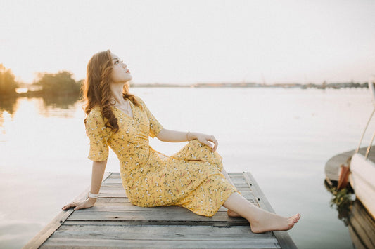 Woman wearing a yellow floral midi dress sitting on a wooden dock by the water, relaxed and confident, representing effortless style after 30