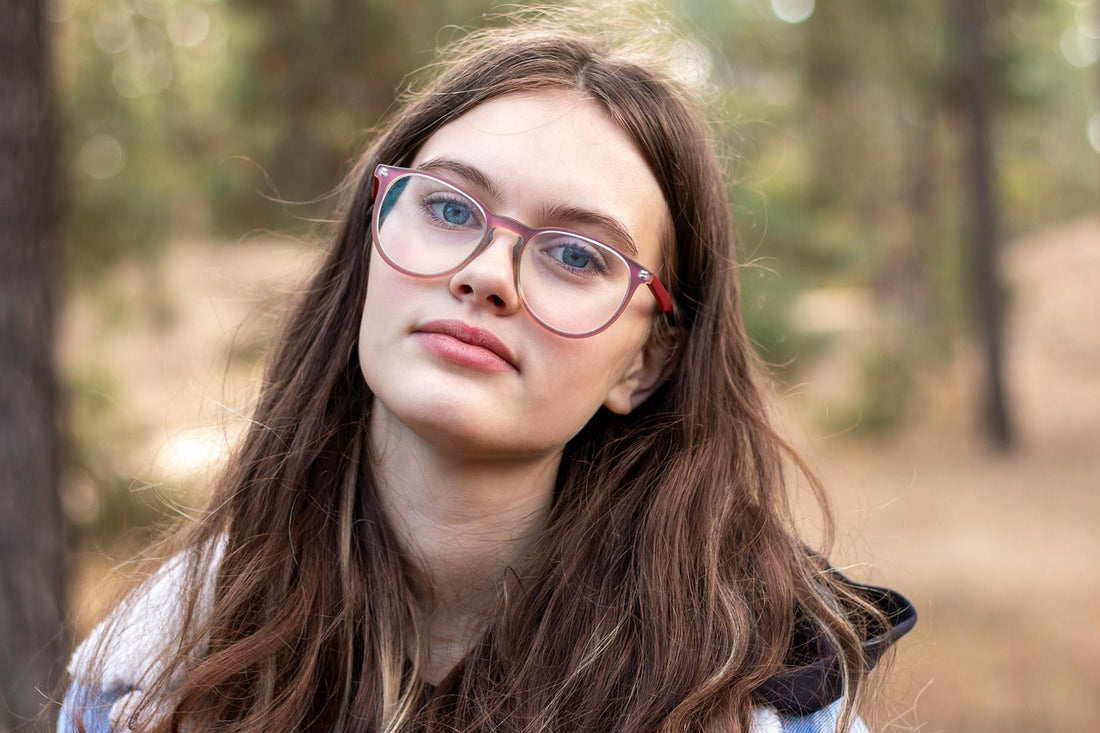 Young woman with long brown hair wearing stylish glasses outdoors, looking at the camera with a neutral expression in a natural forest setting.
