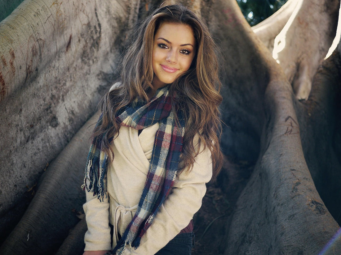 Young woman wearing a beige wrap coat and plaid scarf, posing outdoors against a large tree trunk, showcasing casual fall fashion for women.